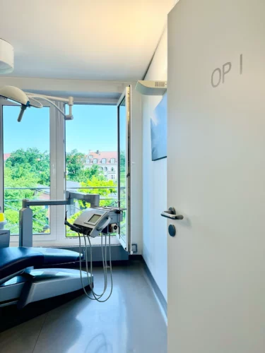 Geöffnete Tür ins helle Behandlungszimmer mit Blick ins Grüne. Auf der Tür steht "OP 1".