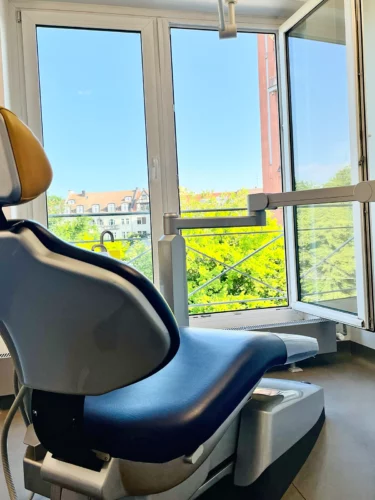 Heller Behandlungsraum mit zahnärztlichem Stuhl am offenen Fenster und Blick ins Grüne.