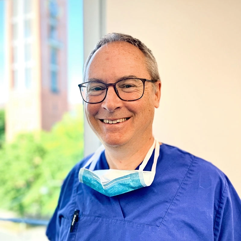 Portraitfoto von Dr. med Stefan Hessenberger in blauem Arzt-Kittel mit umhängenden Mundschutz. Er steht vorm Fenster in seiner Praxis mit einem freundlich entspanntem Lächeln.